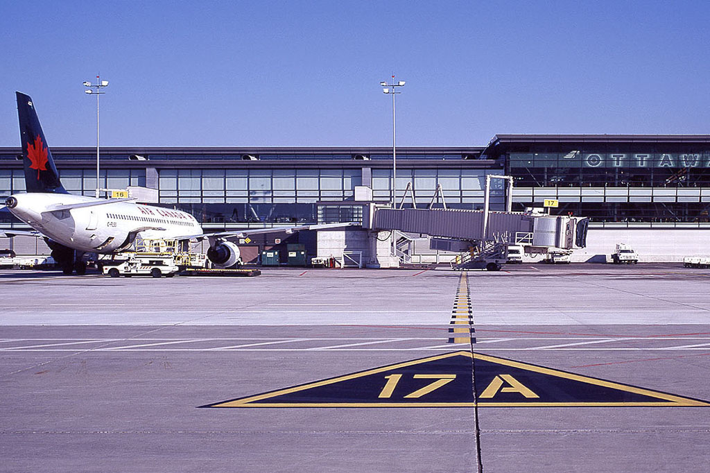 Photo of the Ottawa International Airport from the airside tarmac – a large aircraft is visible on the left side parked at a gate, and the Ottawa wordmark on the glass building façade is clearly visible.